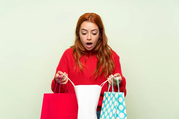 ショッピングバッグの多くを保持する孤立した緑の背景の上にセーターを持つティーンエイジャー赤毛の女の子 — ストック写真