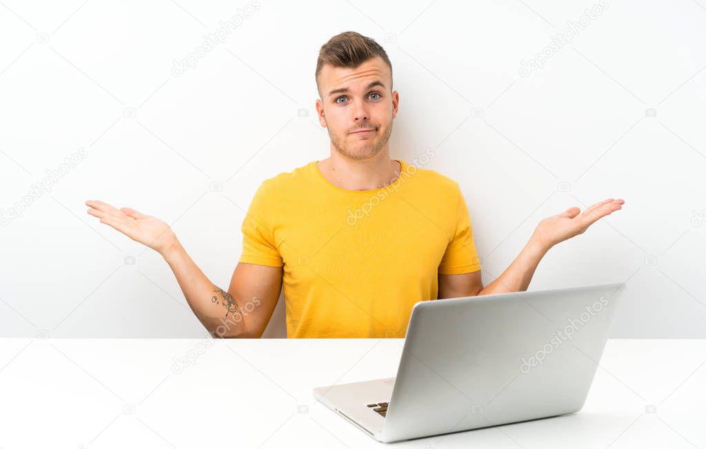 Young blonde man in a table with a laptop having doubts with confuse face expression