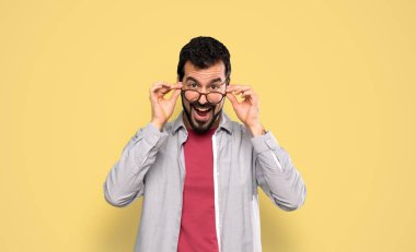 Handsome man with beard with glasses and surprised over isolated yellow background