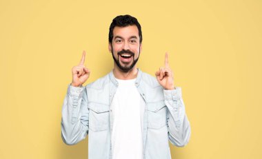 Handsome man with beard pointing up a great idea over isolated yellow background