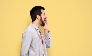 Handsome man with beard shouting with mouth wide open to the lateral over isolated yellow background