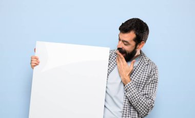 Handsome man with beard holding an empty placard over isolated blue background