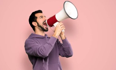Handsome man with sweatshirt shouting through a megaphone over isolated pink background