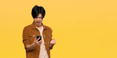 Kahverengi ceketli Asyalı adam zafer pozisyonunda izole edilmiş sarı arka planda.