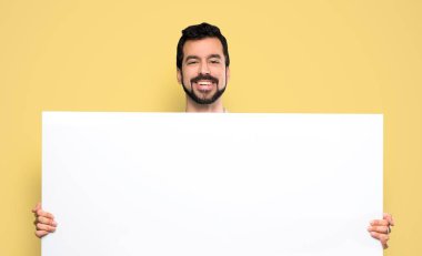 Handsome man with beard holding an empty placard over isolated yellow background