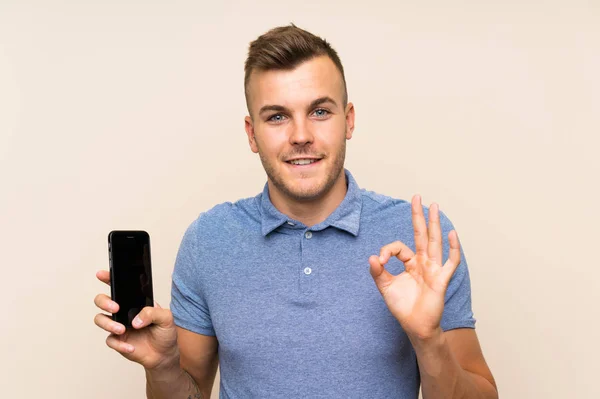 Młody Blondynka Człowiek Pomocą Telefon Komórkowy Pokazując Znak Palcami — Zdjęcie stockowe