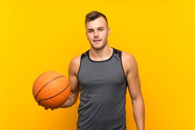 Sarışın ve yakışıklı bir adam, sarı arka planda basket topu tutarken gülümsüyor.