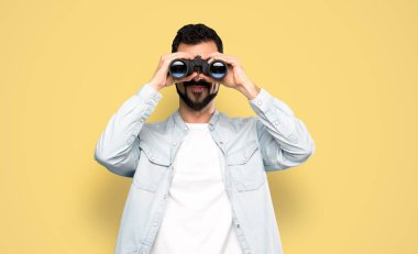 Handsome man with beard with binoculars over isolated yellow background