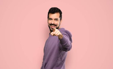Handsome man with sweatshirt points finger at you with a confident expression over isolated pink background