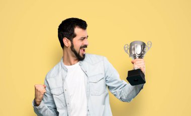 Handsome man with beard holding a trophy over isolated yellow background