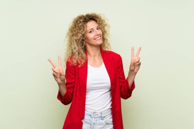 Yeşil arka planda kıvırcık saçlı genç sarışın kadın iki eliyle zafer işareti gösteriyor.