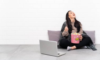 Young Colombian girl holding a bowl of popcorns and showing a film in a laptop celebrating a victory