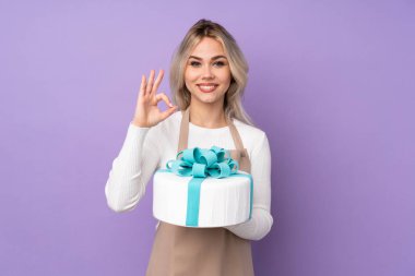 Genç pasta şefi, mor arka planda büyük bir pasta tutuyor. Parmaklarıyla 