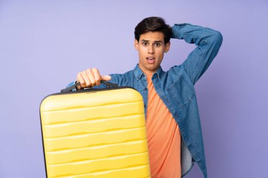 İzole edilmiş mor arka planda seyahat valiziyle gezen ve şaşıran bir adam.