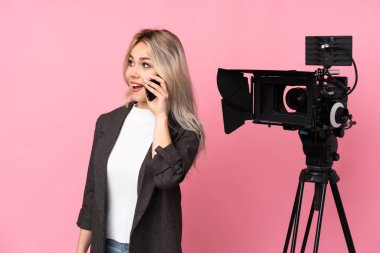 Elinde mikrofon tutan ve pembe arka planda haber sunan muhabir kadın cep telefonuyla birileriyle sohbet ediyor.