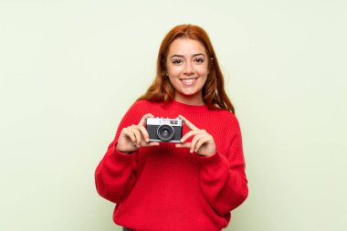 Bir kamera tutan izole yeşil arka plan üzerinde kazak ile genç redhead kız