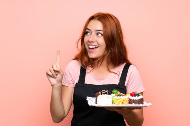 Genç kızıl saçlı kız pembe arka planda bir sürü farklı mini kek tutuyor. Çözümü bulmak için parmağını kaldırıyor.