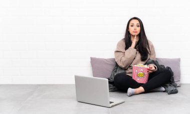 Young Colombian girl holding a bowl of popcorns and showing a film in a laptop keeps palm together. Person asks for something