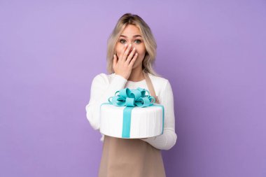 Genç pasta şefi, mor arka planda büyük bir pasta tutuyor ve yüzünde sürpriz bir ifade var.