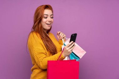 Mor arka planda kalan kızıl saçlı genç kız elinde alışveriş torbalarıyla bir arkadaşına cep telefonuyla bir mesaj yazıyor.