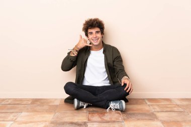 Genç beyaz bir adam yerde oturuyor ve telefon jesti yapıyor. Bana geri dönüş işareti ver.