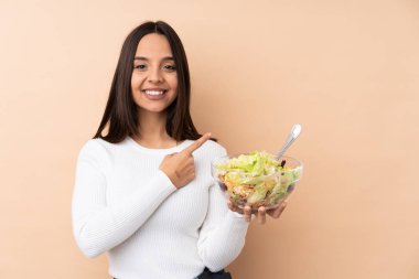 Genç esmer kız izole edilmiş arka planda salata tutuyor. Ürünü sunmak için yan tarafı işaret ediyor.