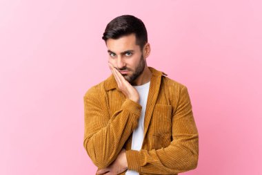 Sakallı genç yakışıklı adam pembe arka planda kadife ceket giyiyor mutsuz ve hayal kırıklığına uğramış.
