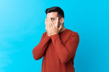 Sakallı, mavi arka planda genç ve yakışıklı bir adam. Gözleri kapatıyor ve parmaklarından bakıyor.