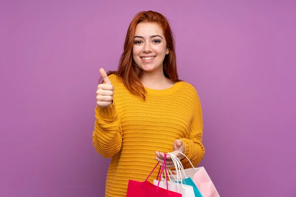 赤毛ティーン女の子上の隔離された紫色の背景保持ショッピングバッグととともに親指アップ — ストック写真