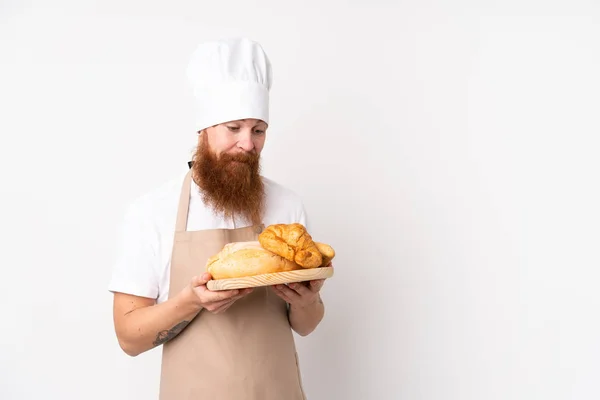 Uomo dai capelli rossi in uniforme da chef. Fornaio maschio che tiene un tavolo con diversi tipi di pane — Foto Stock