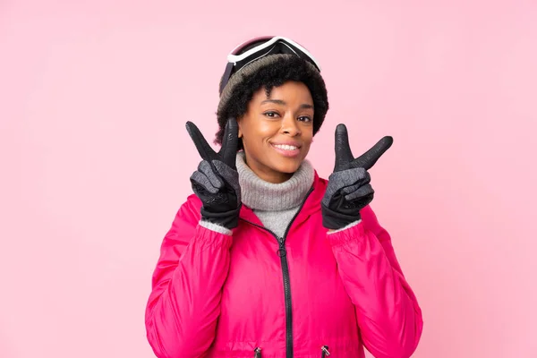 非洲裔美国滑雪者 戴着雪板眼镜 带着淡淡的粉色背景 微笑着 并展示出胜利的标志 — 图库照片