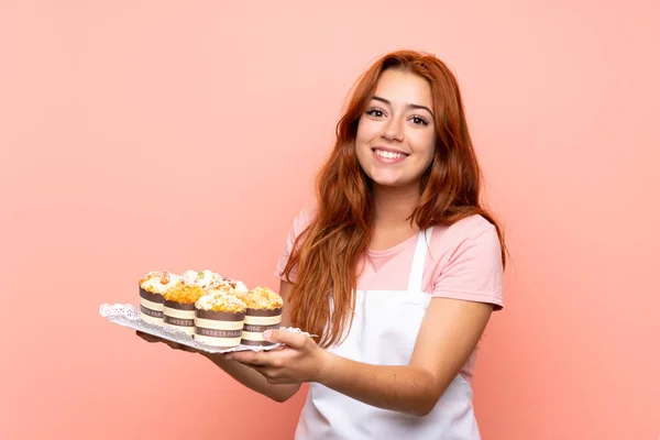 Adolescente rousse fille tenant beaucoup de différents mini gâteaux sur fond rose isolé — Photo