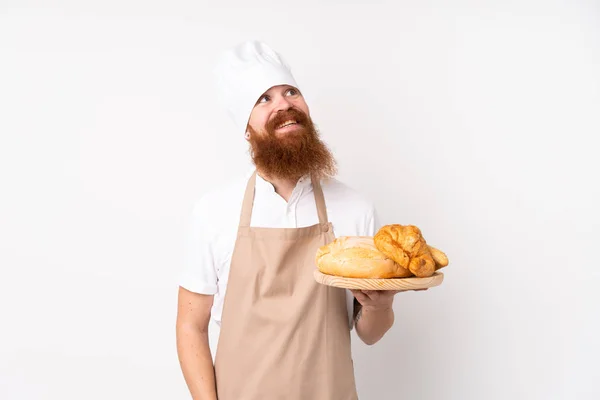 シェフの制服を着た赤毛の男 男のパン屋がテーブルを持っているいくつかのパンを見上げながら笑顔 — ストック写真