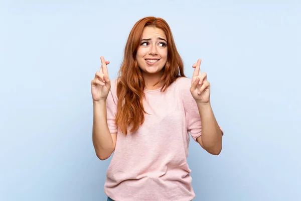 Teenager redhead girl over isolated blue background with fingers crossing and wishing the best