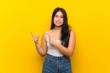 Young teenager Asian girl over isolated yellow background frightened and pointing to the side