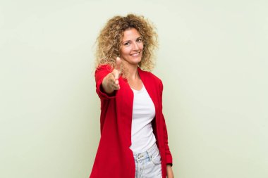 Sarışın, kıvırcık saçlı, yalıtılmış yeşil arka planı olan genç bir kadın iyi bir anlaşma için el sıkışıyor.