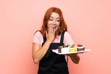 Genç redhead kız sürpriz ve şok yüz ifadesi ile izole pembe arka plan üzerinde farklı mini kek çok tutan