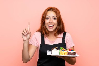 İzole edilmiş pembe arka plan üzerinde bir sürü farklı mini kek tutan genç kızıl kız harika bir fikre işaret ediyor.