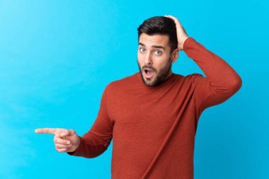 Sakallı, mavi arka planda genç ve yakışıklı bir adam. Şaşırmış ve işaret ediyor.