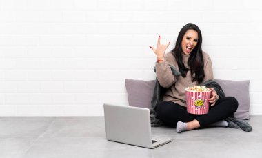 Young Colombian girl holding a bowl of popcorns and showing a film in a laptop making rock gesture