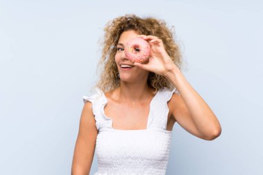 İzole mavi arka plan üzerinde bir donut tutan kıvırcık saçlı genç sarışın kadın