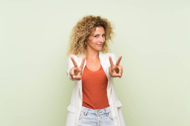Yeşil arka planda kıvırcık saçlı genç sarışın kadın gülümsüyor ve zafer işareti gösteriyor.