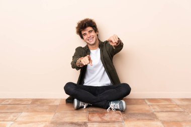 Yerde oturan genç beyaz adam gülümserken seni işaret ediyor.