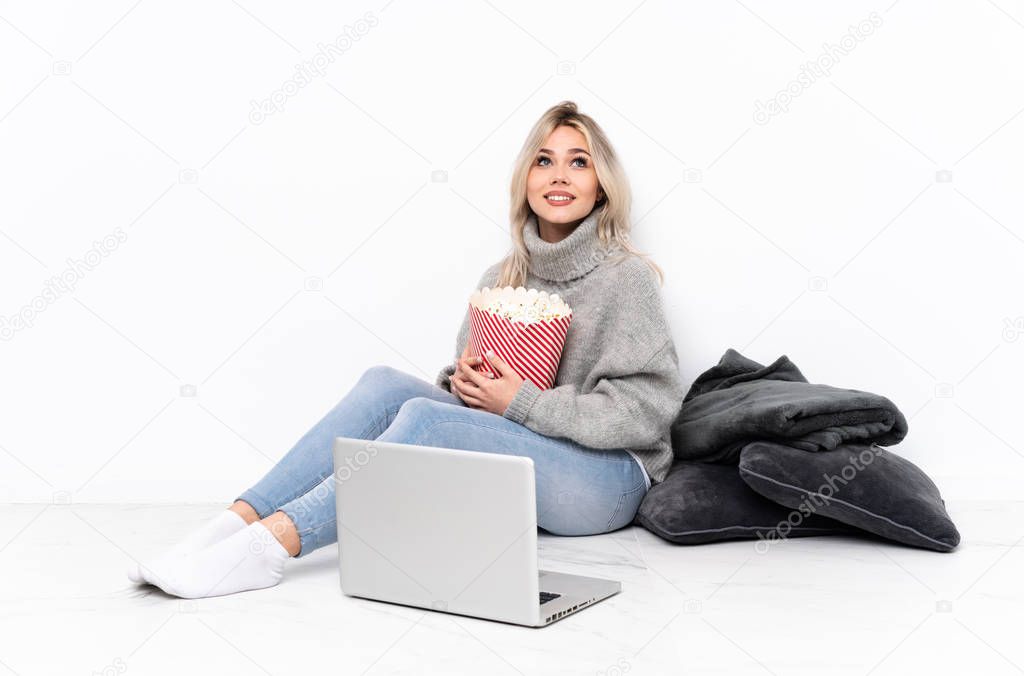 Teenager blonde girl eating popcorn while watching a movie on the laptop looking up while smiling