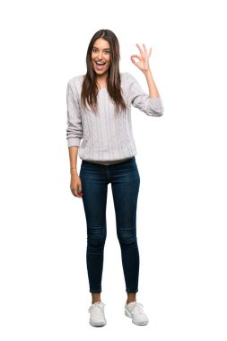A full-length shot of a Young hispanic brunette woman surprised and showing ok sign over isolated white background