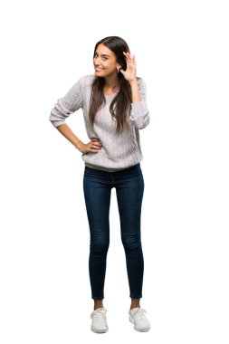 A full-length shot of a Young hispanic brunette woman listening to something by putting hand on the ear over isolated white background