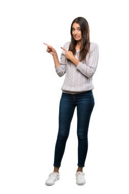 A full-length shot of a Young hispanic brunette woman frightened and pointing to the side over isolated white background
