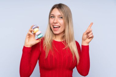 Soyutlanmış mavi arka planı olan genç sarışın kadın elinde renkli Fransız badem kurabiyeleriyle harika bir fikir sunuyor.