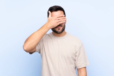 Sakallı, genç, yakışıklı, soyutlanmış, mavi arka planda, gözleri elle kaplanmış.