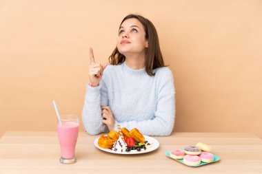 Bej arka planda izole edilmiş waffle yiyen genç kız yukarı bakıyor ve şaşırıyor.